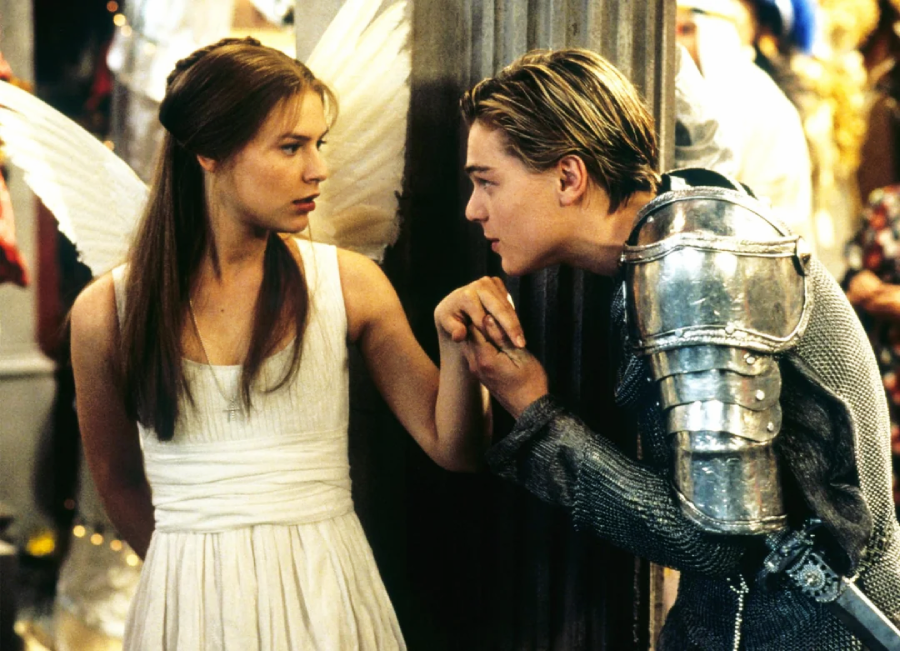 A young woman in a white dress with angel wings stands as a young man in silver armor gently kisses her hand. They look at each other intensely in a dramatic, costumed setting.