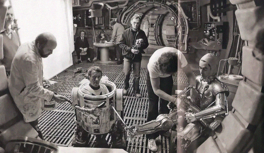 Behind-the-scenes view of "Star Wars" set, showing crew members assisting actors inside R2-D2 and C-3PO costumes, with film equipment and set pieces visible in the background. Black and white photo.