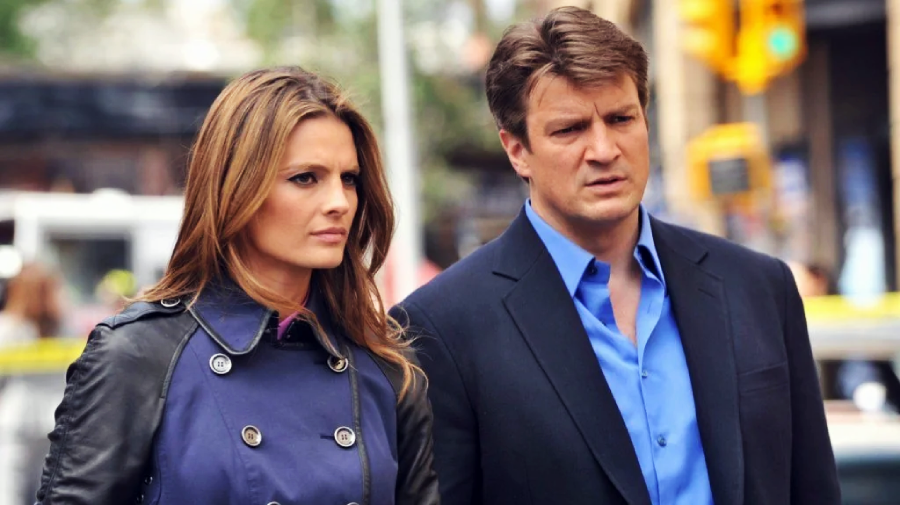 A woman in a blue trench coat and a man in a blue shirt and dark jacket stand side by side outdoors, both looking serious. The background is slightly blurred with yellow caution tape and city elements visible.