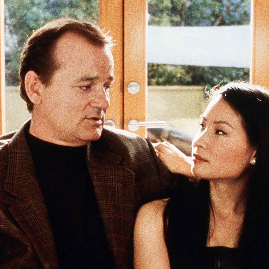 A man in a brown plaid jacket and black shirt sits closely with a woman in a black dress. They are indoors, and the woman has her arm on the man's shoulder as they look at each other.