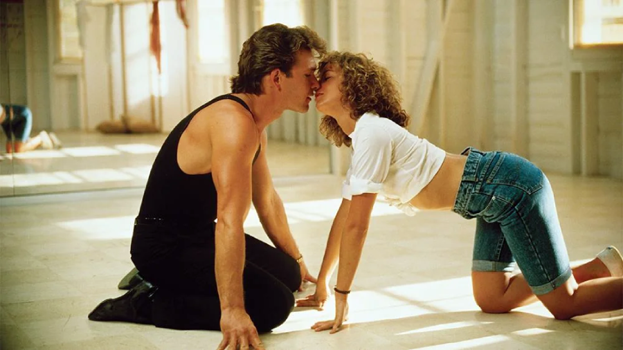 A man in a black tank top and a woman in a white crop top and denim shorts are kneeling on the floor, facing each other closely, about to kiss in a brightly lit dance studio.