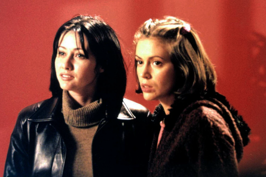 Two young women with serious expressions stand close together against a red background. One has dark hair and wears a leather jacket over a sweater, while the other has lighter hair and wears a dark sweater with a hair clip.