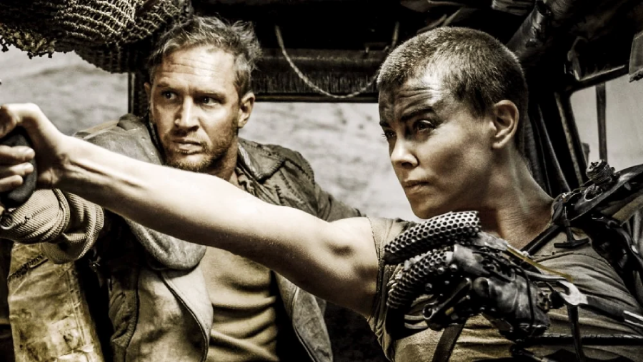A man and a woman with dirt-smeared faces sit in a rugged vehicle. The woman, with a buzz cut, aims a gun, while the man looks intently in the same direction, ready for action in a post-apocalyptic setting.