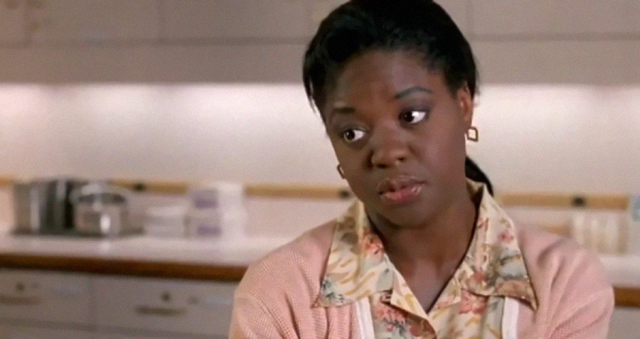 A woman wearing a light floral blouse and pink cardigan looks to the side with a slightly skeptical or uncertain expression. She is in a kitchen or break room with a blurred background of cabinets and counters.