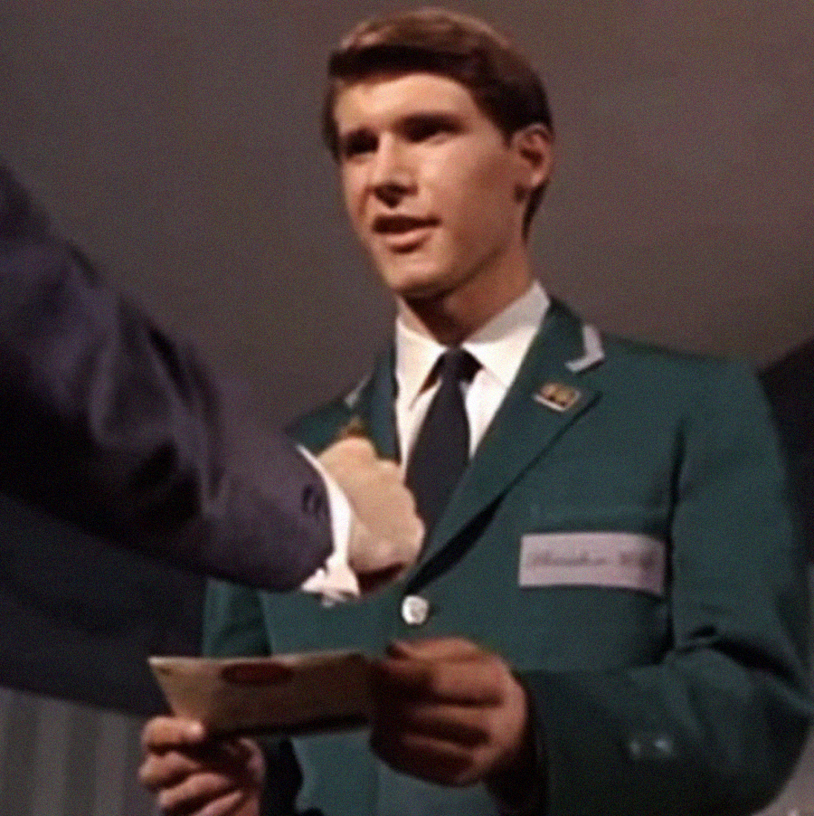 A young man in a green blazer and tie stands indoors, holding an envelope while another person’s arm reaches toward him, possibly in a gesture of congratulations or handing over the item.