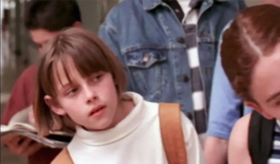 A young girl with straight brown hair and bangs, wearing a white turtleneck and brown backpack, looks skeptically to her side. Other children are visible in the blurry background.