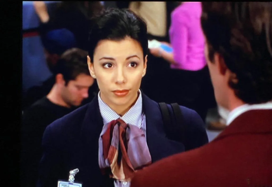 A woman with dark hair tied back, wearing a suit and a large bow tie, looks at a man in a maroon jacket. She has a neutral expression, and people are blurred in the background.