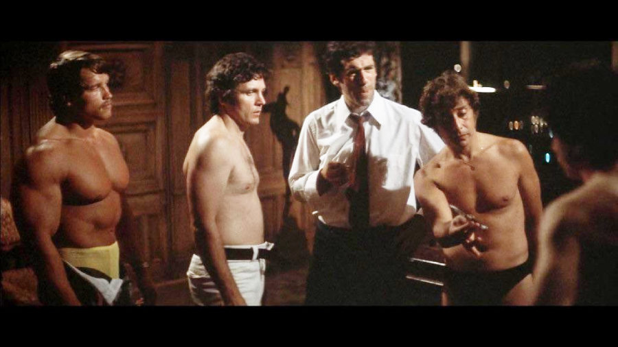 Four men stand indoors, three of them shirtless and wearing only underwear or shorts, while the fourth wears a shirt and tie. One shirtless man is gesturing toward another man, and all appear to be engaged in conversation.