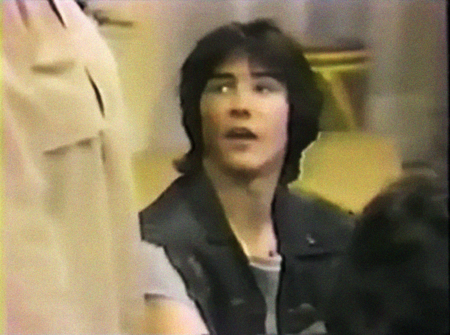 A young man with medium-length dark hair and a black jacket looks upward, mouth slightly open, in a grainy, vintage-style image.