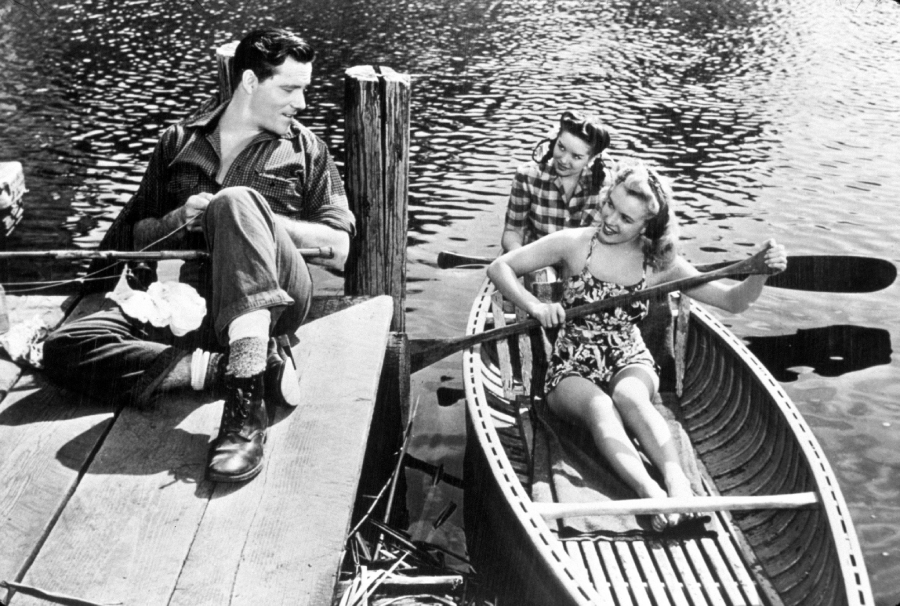 A man sits on a dock smiling at two women in a small rowboat on the water. One woman is rowing while the other sits behind her, both looking up at the man. The scene appears to be from the mid-20th century.