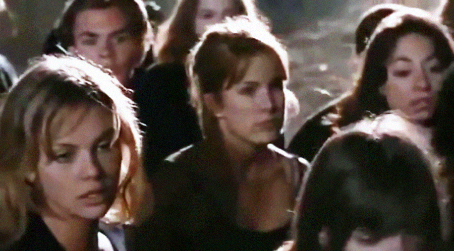 A group of people stands close together outdoors at night, looking serious or concerned. The scene is dimly lit and slightly blurry.