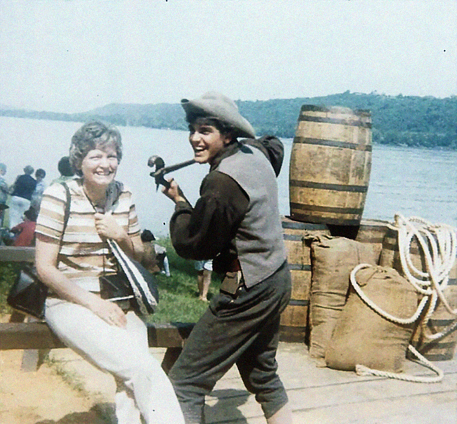 A woman sits smiling on a bench near a river while a man in old-fashioned clothes and a hat poses playfully with a cane over his shoulder. Barrels, sacks, and ropes are stacked in the background, with hills and water visible.