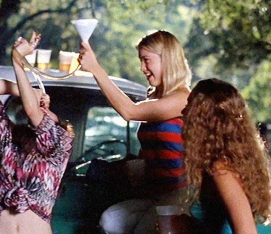 Three young women are outside near a car; one uses a beer bong while another holds it up and laughs. The third woman with long curly hair stands nearby. There are cups of beer on the car roof, and trees are visible in the background.
