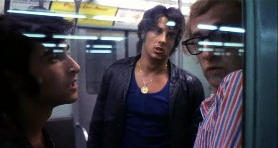 Three men stand closely together inside a subway car. Two of them, in darker clothing, face the third man, who wears glasses and a striped shirt. Fluorescent lights and metal surfaces are visible in the background.