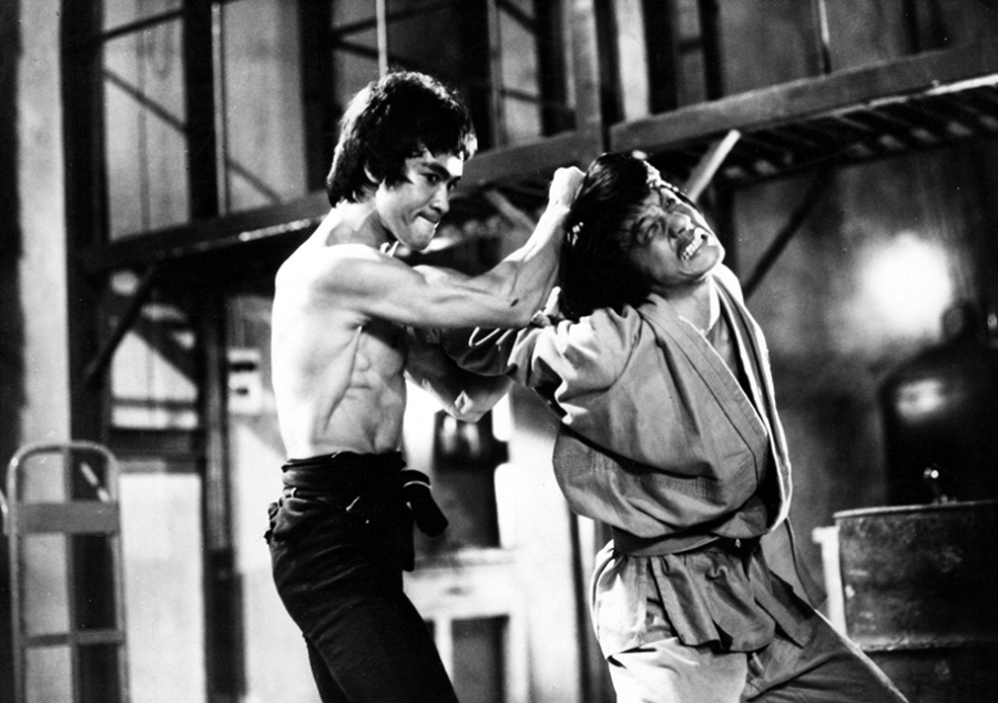 A shirtless martial artist grabs another man by the hair during an intense fight scene in an industrial setting, both displaying dramatic expressions and dynamic movement.