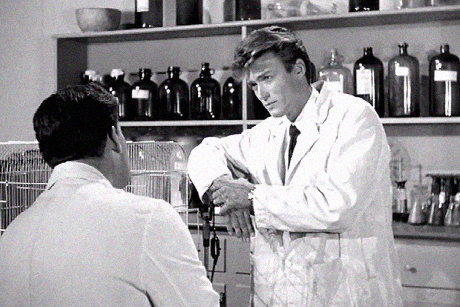 Two men in lab coats talk in a laboratory, with shelves of bottles and scientific equipment behind them. One man leans on a table, facing the other, who sits near a wire cage. The scene is in black and white.