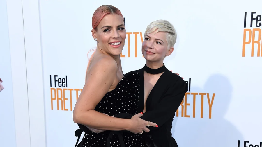 Two women pose together on a red carpet in front of an "I Feel Pretty" backdrop. One wears a black polka dot dress with pink hair, the other has a short blonde haircut and wears a black outfit. They smile and embrace.