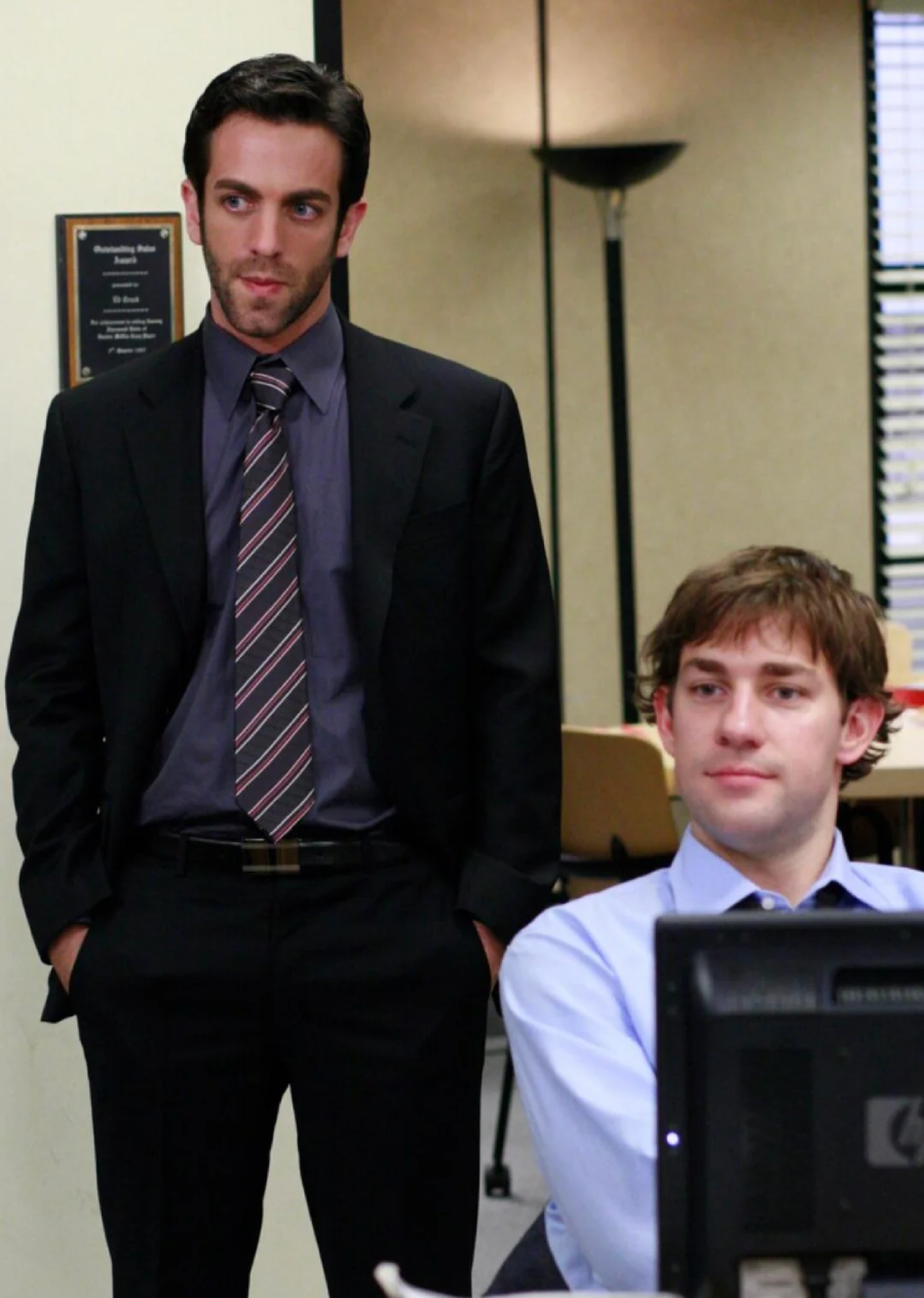 Two men in an office setting, one standing in a black suit and striped tie with hands in his pockets, the other sitting at a desk with a computer, wearing a light blue shirt. Both appear to be looking at something off-camera.