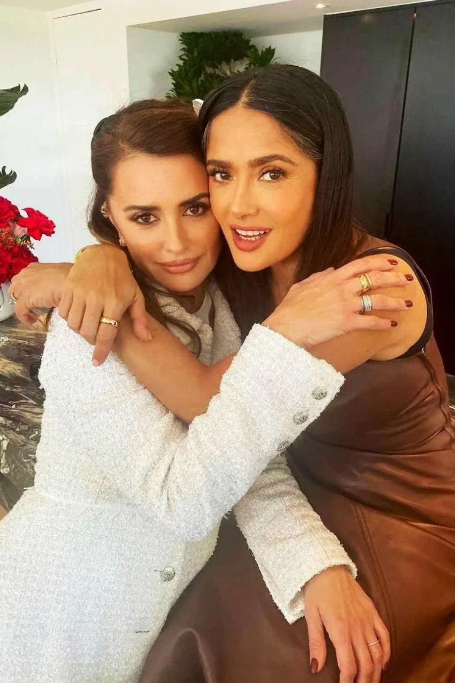 Two women are sitting closely together, smiling, and hugging each other. One is wearing a white outfit and the other a brown dress. There are red flowers and a plant in the background.