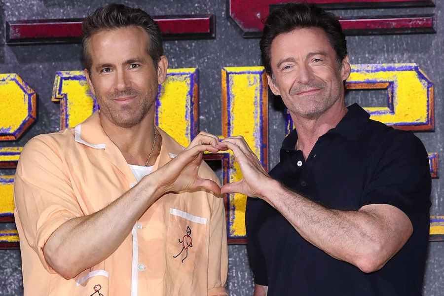 Two men stand side by side, smiling, and forming a heart shape with their hands. One wears a peach shirt over a white T-shirt; the other wears a black polo. They are in front of a wall with large, colorful letters.