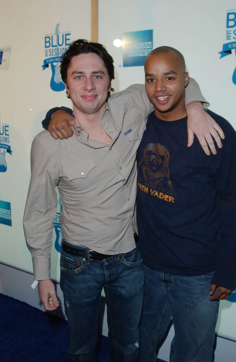Two men stand close together and smile at the camera on a blue carpet. One man has his arm around the other. Both are casually dressed in jeans and shirts, and there are event banners in the background.