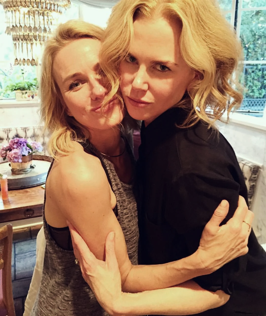 Two women with blonde hair are hugging each other and smiling indoors. One is wearing a sleeveless gray top, and the other is in a black shirt. The background shows a dining area with flowers and a chandelier.