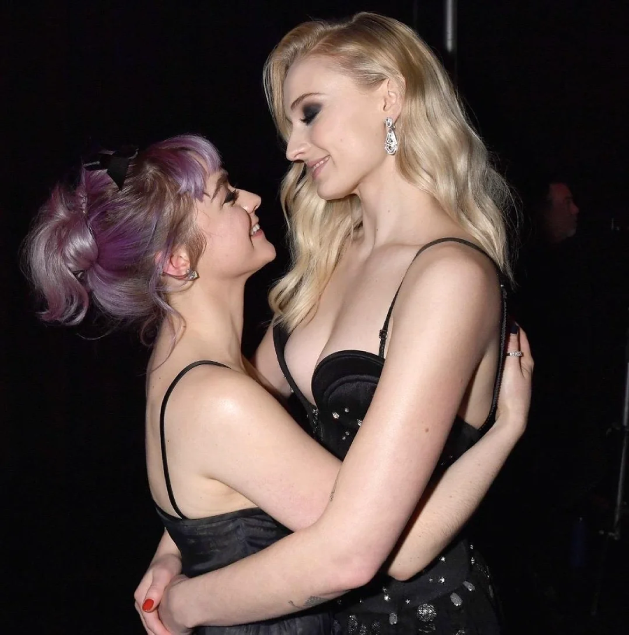 Two women in black dresses embrace and smile at each other affectionately, with one having purple hair styled up and the other with long blonde hair. The background is dark, drawing focus to their joyful expressions.