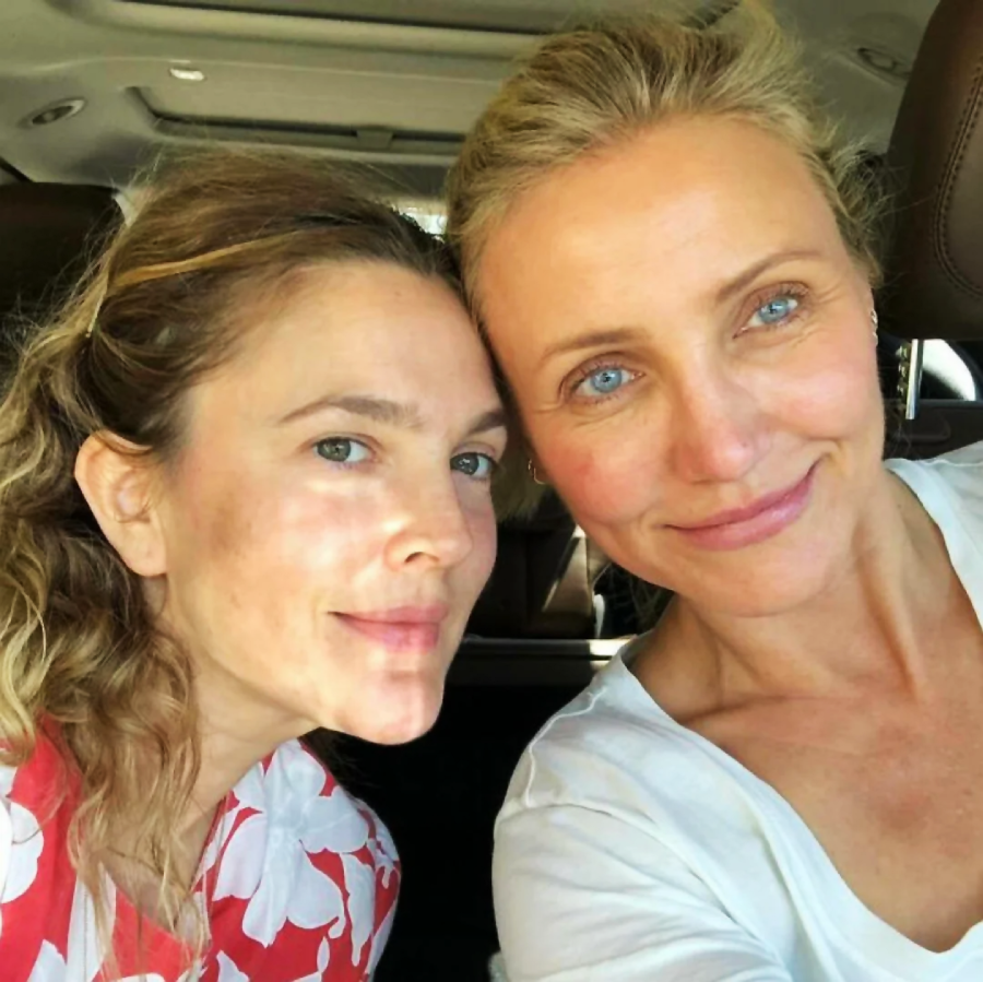 Two women sitting close together in a car, smiling softly at the camera. One has wavy hair and wears a red and white top; the other has light eyes, blonde hair, and wears a white shirt. Sunlight filters onto their faces.