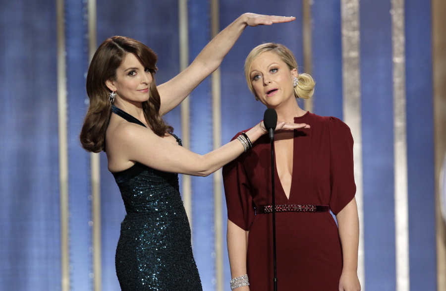Two women stand on stage; one in a black sequin dress gestures with her hands above and below the other’s head, who wears a deep red dress and stands at a microphone, both appearing playful and expressive.