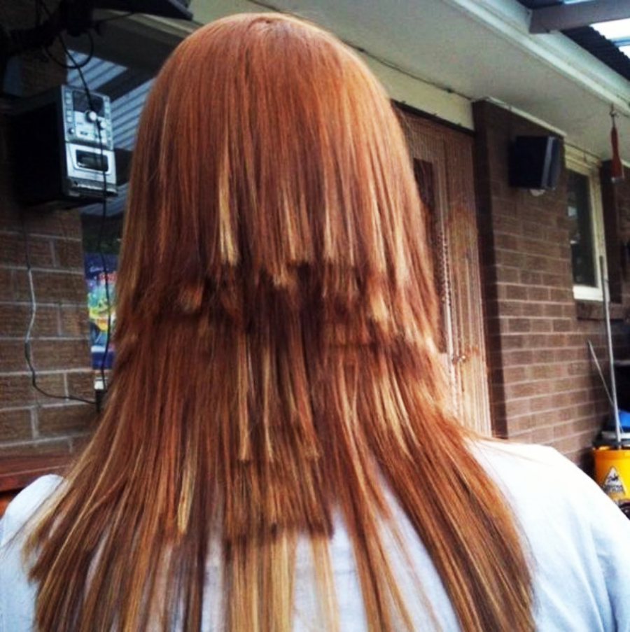 A person with long, straight reddish-brown hair cut into uneven, stair-like layers, standing outside near a brick wall and a door.