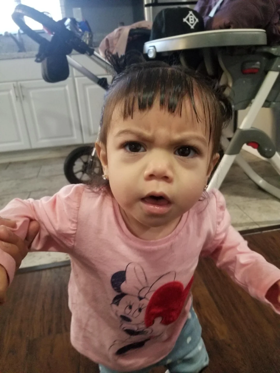 A young toddler with short bangs and pigtails wears a pink Minnie Mouse shirt, looking up with a serious, confused expression indoors near a stroller and high chair.