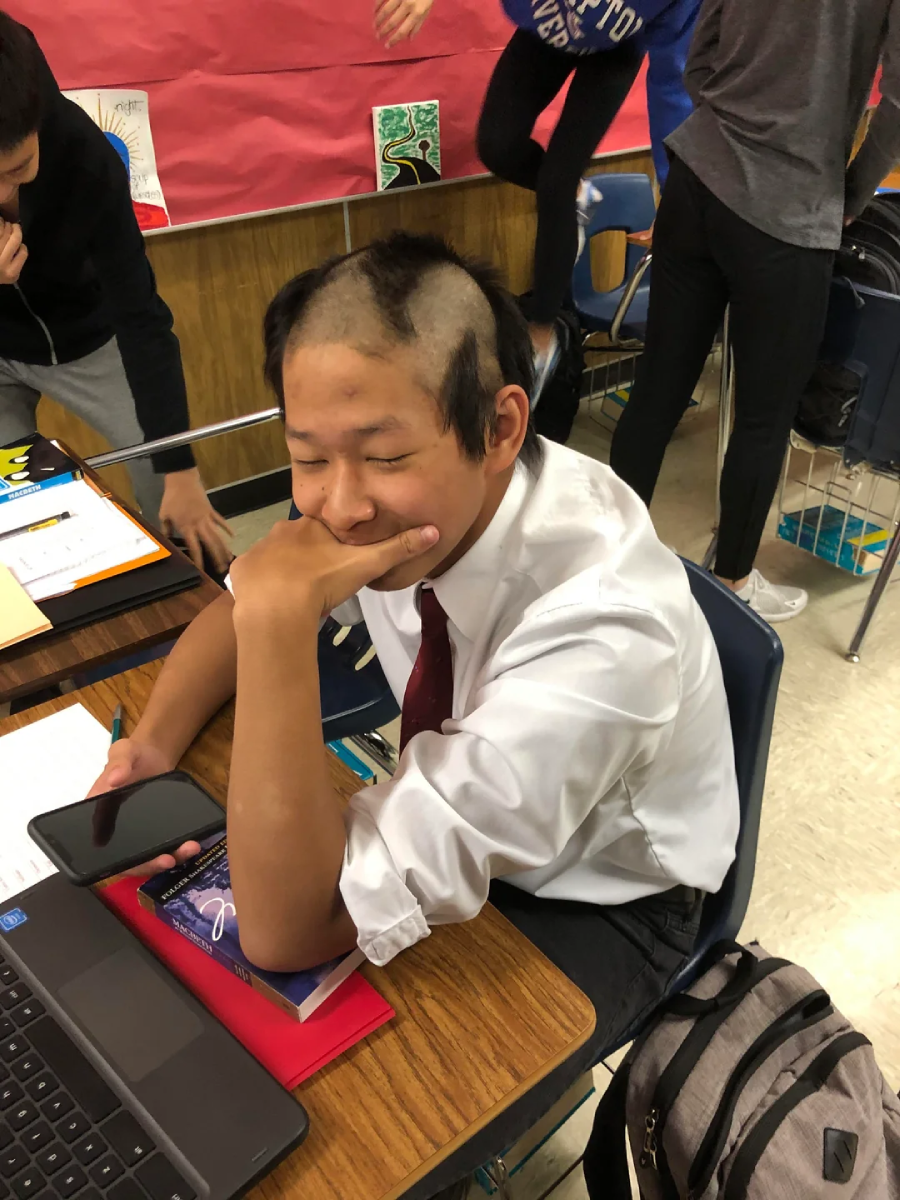 A student with a patchy, uneven haircut sits at a classroom desk, resting his chin on his hand and smiling with his eyes closed. He wears a white shirt and red tie, with a laptop and school supplies on the desk.