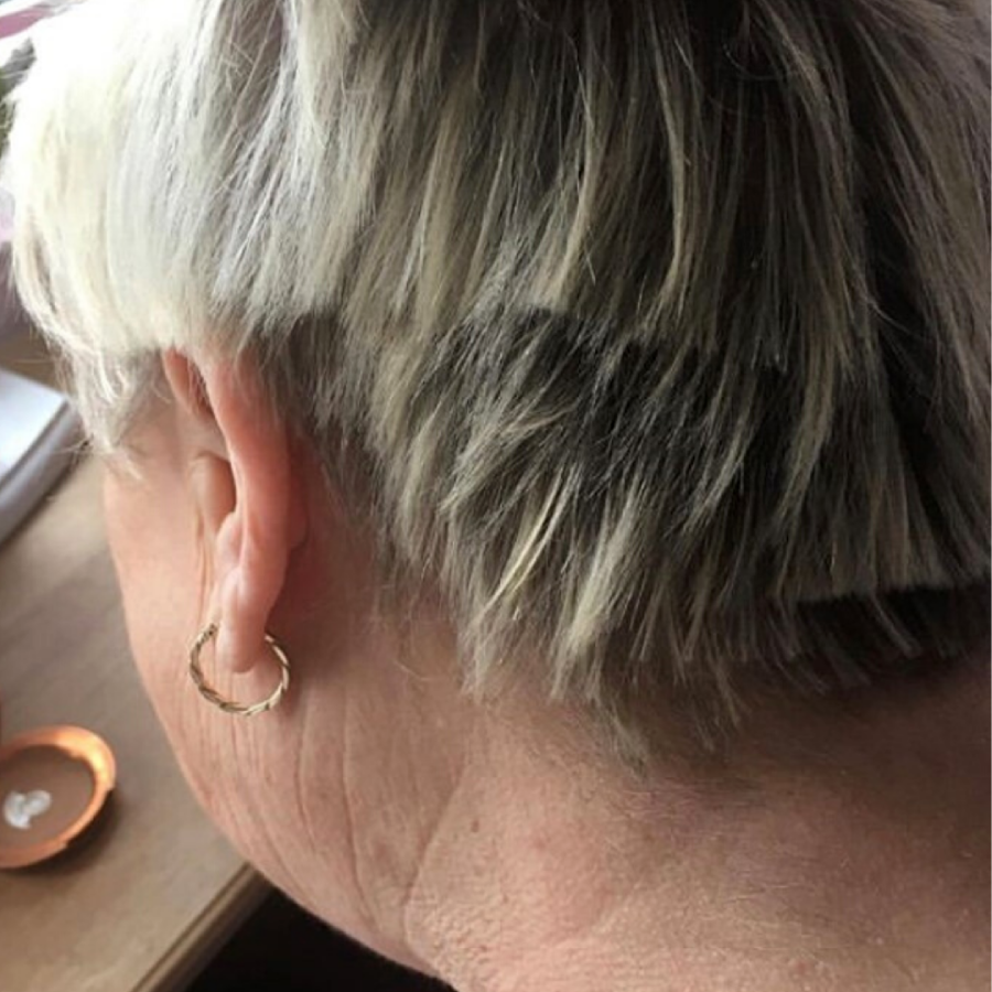 A close-up of the back of a person's head with short, layered gray and blonde hair, showing a gold hoop earring in one ear. The person is seated near a table with a small copper dish on it.