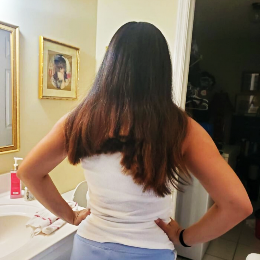 A woman stands in a bathroom facing away, showing unevenly cut long brown hair. She wears a white tank top and light blue pants, with her hands on her hips. A framed photo, towels, and soap are visible on the sink.