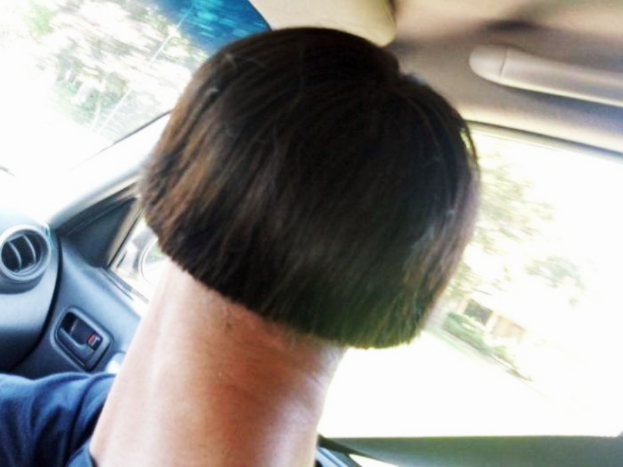A person sitting in a car with the back of their head visible, showing a bowl-shaped haircut that creates an unusual visual effect with their neck.