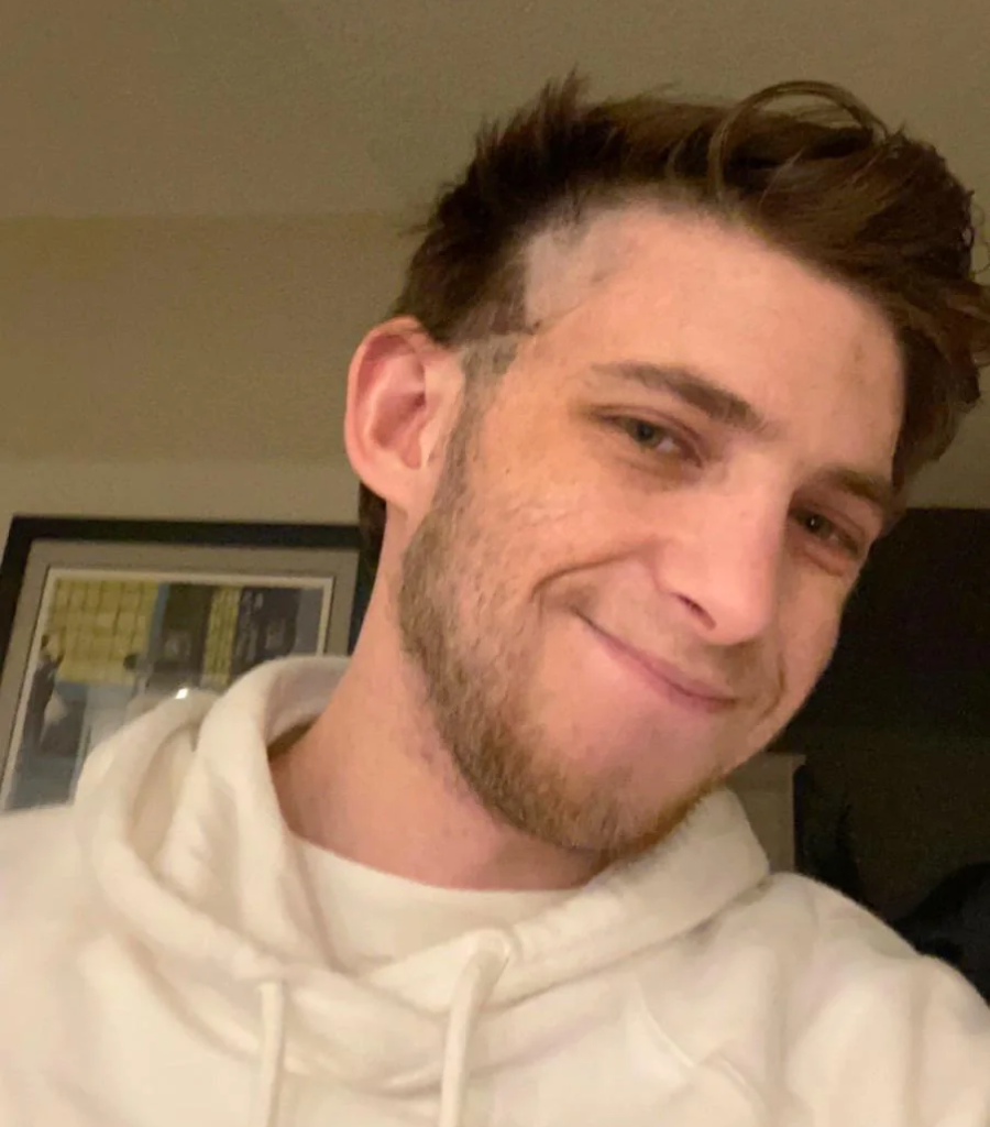 A young man with short brown hair, patchy shaved areas around his ear, slight beard, and a white hoodie smiles at the camera indoors, with a framed picture on the wall behind him.