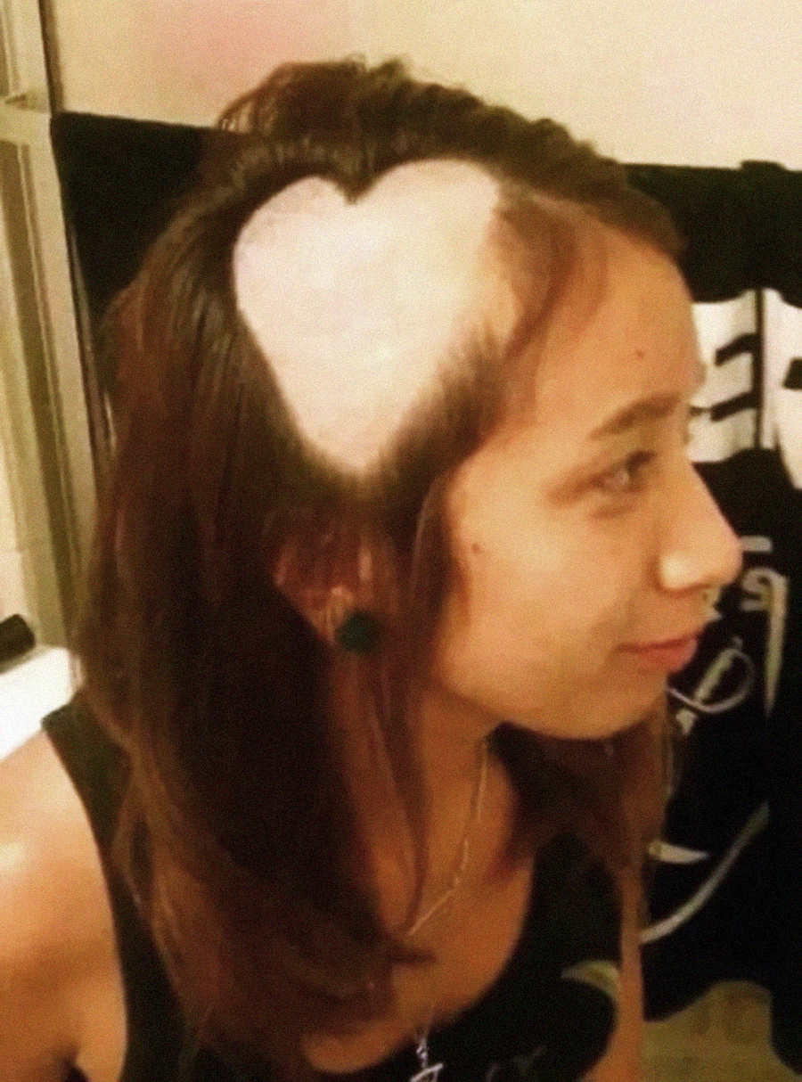 A woman with long brown hair has a heart-shaped section shaved on the side of her head, revealing her scalp in the shape of a heart. She is smiling and facing right.