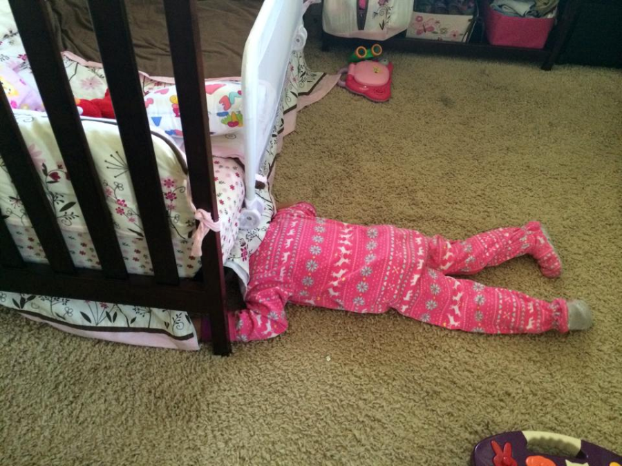 A young child in pink patterned pajamas lies face down on a carpet, half under a crib, with only their legs and lower torso visible. The room has a soft brown carpet and children's items nearby.