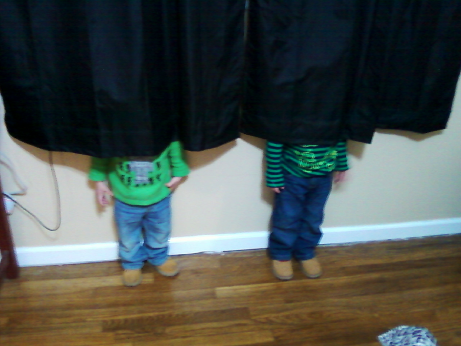 Two children stand side by side with their heads hidden behind black curtains, only their bodies in green shirts, jeans, and tan shoes visible against a tan wall and wooden floor.