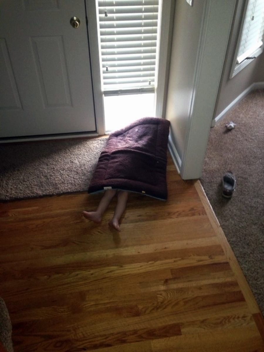 A child is lying on the floor partly outside a door, covered by a mat so only their bare legs and feet are visible. Blinds are partially open, letting in sunlight. A shoe and a sock are nearby on the carpet.