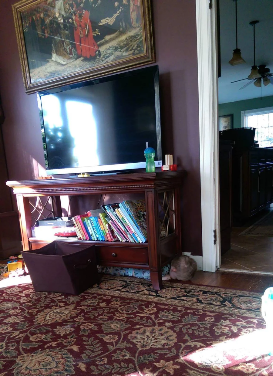 A living room with a TV on a wooden stand, children's books on a shelf, a toy bin on the floor, a green sippy cup on the stand, and sunlight streaming in from a window. A cat is curled up beside the stand.