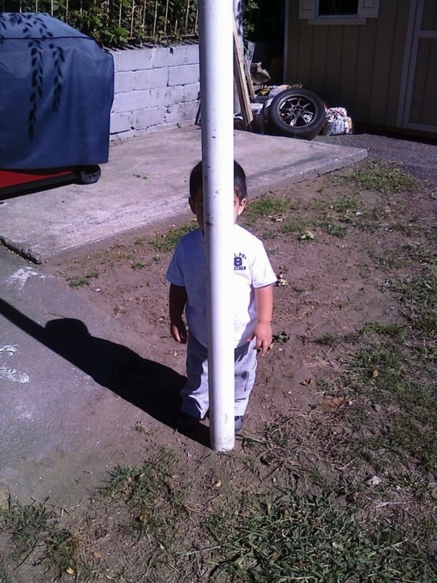 A child stands behind a white pole, which perfectly aligns to hide the middle of their face and body, creating a humorous visual effect. The scene is outdoors, near a patio with various objects in the background.