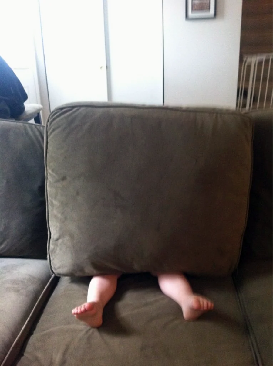 A child hides behind a large couch cushion with only their bare legs and feet visible sticking out from underneath.