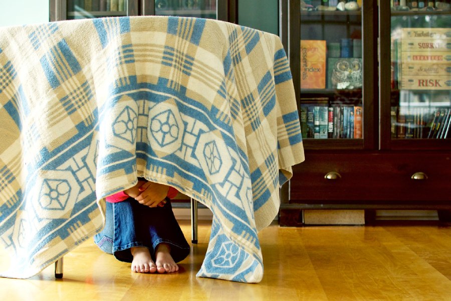 A child sits barefoot under a table covered with a blue and beige patterned blanket, with only their hands and feet visible. Bookshelves filled with books are in the background.