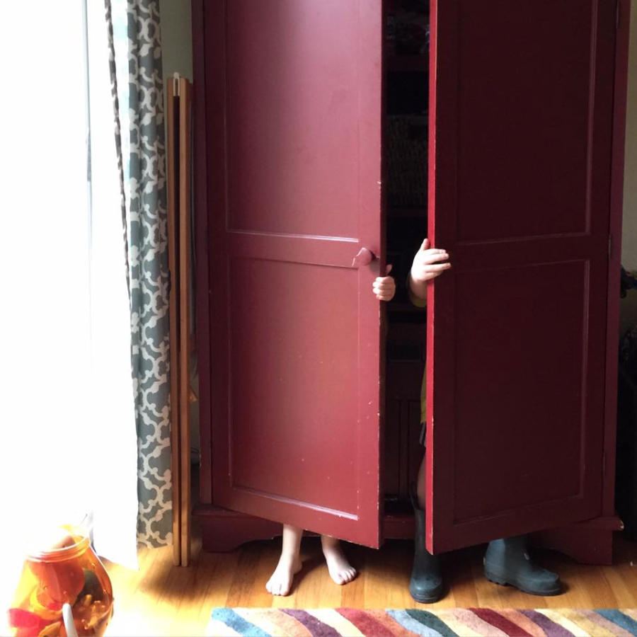Two children are hiding inside a large red wardrobe; only their feet and hands are visible as they peek out from behind the partially opened doors. A colorful rug and a fishbowl are on the floor nearby.