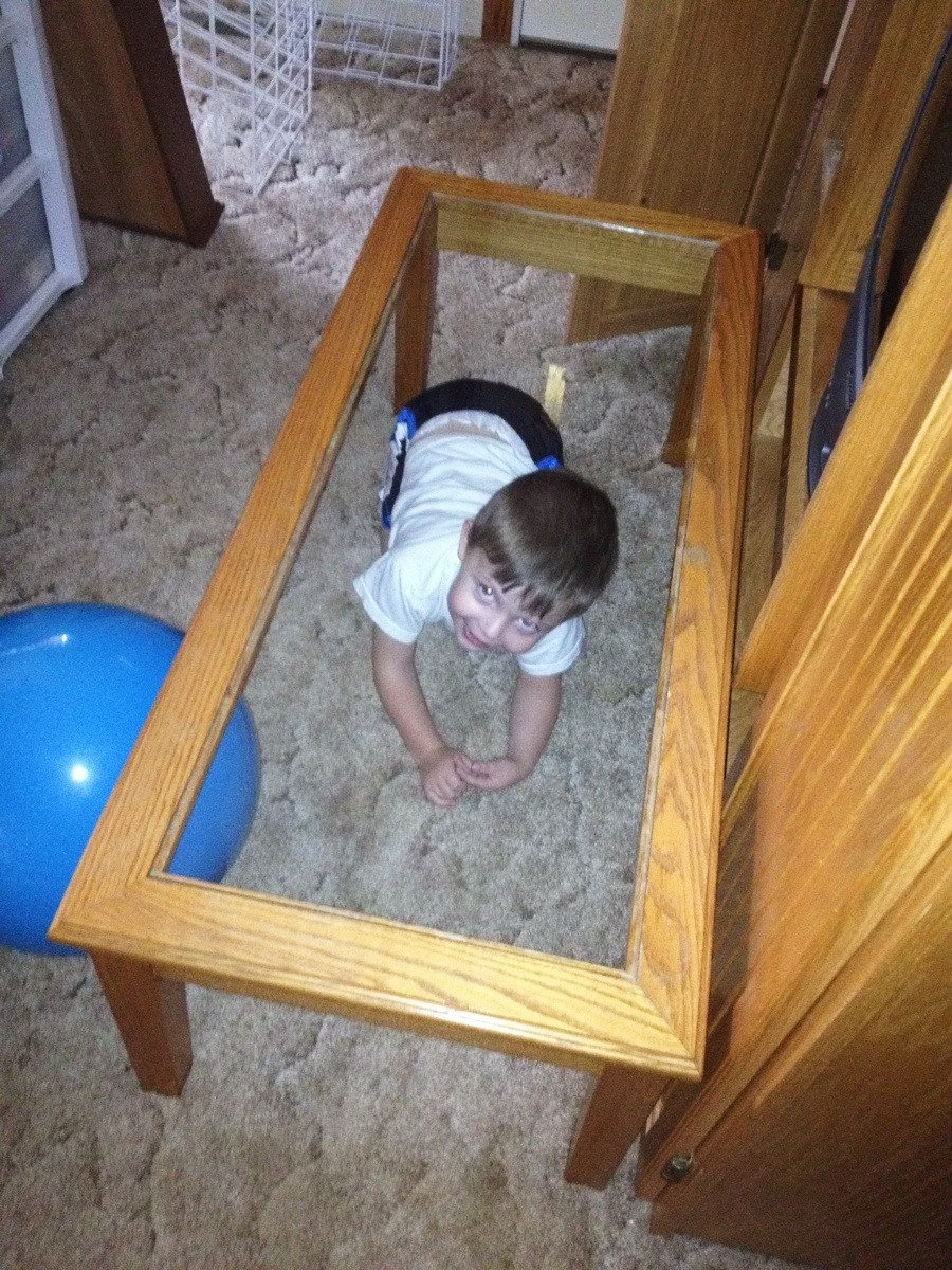A young child is lying on the carpeted floor beneath a rectangular wooden coffee table with a glass top, looking up and smiling. A large blue ball is nearby.