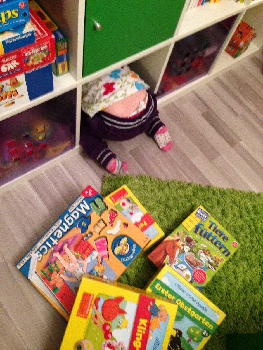 A small child with floral pants and striped socks is crawling headfirst into a cubby shelf. Several colorful children's books are scattered on the floor nearby. The scene is playful and indoors.