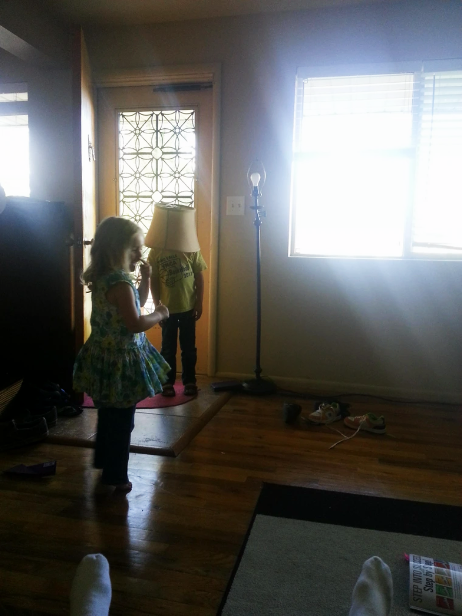 Two children stand in a sunlit living room. One child faces the camera, while the other stands near the door with a lampshade covering their head. Shoes are scattered on the floor, and sunlight streams through a window.
