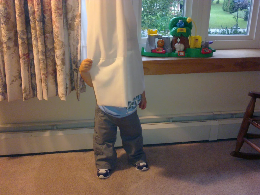 A small child stands behind a curtain with their head completely covered, leaving only their lower body and legs visible. There are toys and a window on the wooden sill in the background.