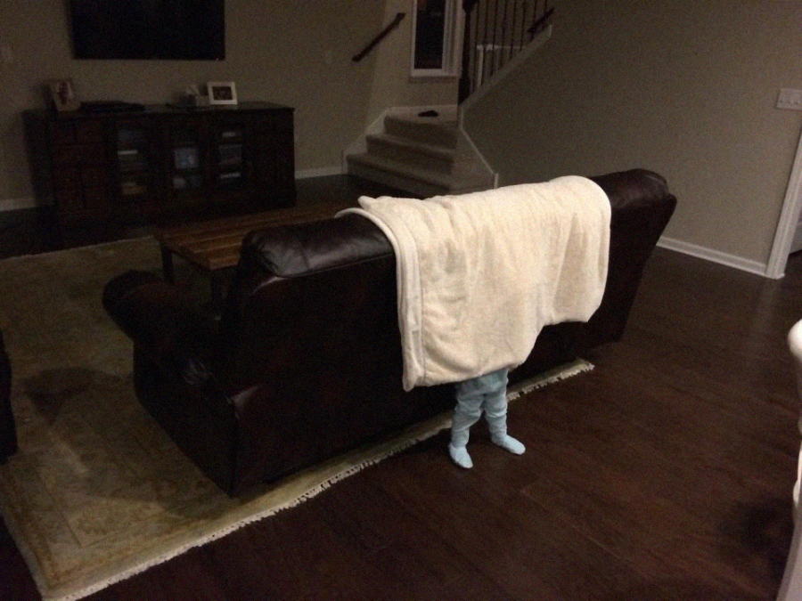 A small child, mostly hidden behind a dark brown couch draped with a white blanket, stands with only their legs visible in a dimly lit living room with wooden floors.
